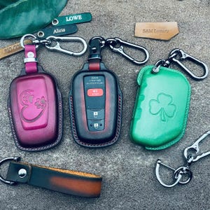 May include: Three leather key fob covers with metal carabiner clips. One is magenta with a heart and paw print, one is black and red with a visible car key remote, and one is green with a shamrock. A leather strap and small name tags are included.