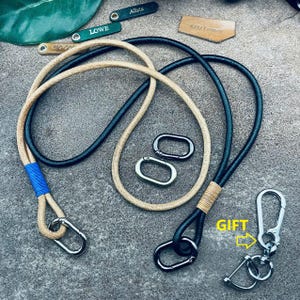 May include: Two leather lanyards, one black and one beige, with blue trim, and two silver swivel clips. The lanyards are tied together with a brown cord. A silver keychain with a swivel clip is included as a gift.