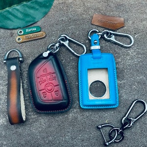 May include: A collection of leather keychains and key fobs in various colors. Includes a black and red key fob, a blue key fob, and a brown leather keychain. Also includes small tags with the names "Karina" and "Big Goldy".