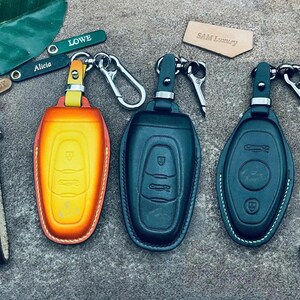 Könnte beinhalten: Leder-Schlüsselanhänger in verschiedenen Farben, darunter leuchtendes Orange und Gelb sowie ein tiefes Teal. Jeder Anhänger hat einen silbernen Schlüsselring und ein geprägtes Autodesign auf der Vorderseite. Die Abbildung zeigt auch Schlüsselanhänger mit Namen.