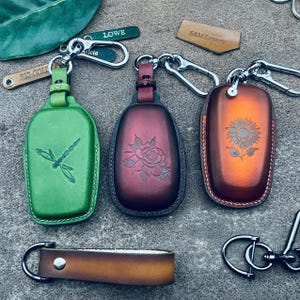 Op de afbeelding: Drie lederen sleutelhangerhoezen in groen, bordeauxrood en oranje verloop, elk voorzien van reli&euml;fmotieven van een libel, een roos en een zonnebloem. Elke hoes bevat een zilverkleurige metalen karabijnhaak en een bijpassende lederen riem.