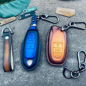 May include: Two leather keychains with key fob covers. One is blue and the other is brown. Both have three buttons on the front. The keychains have metal rings and hooks.