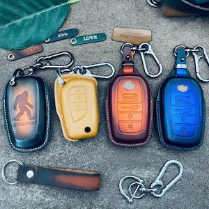 May include: A collection of leather keychains and key fobs in various colors. The key fobs include a yellow, brown, and blue design. One keychain features a Bigfoot graphic. Other keychains have names like "Alicia" and "Lowe".