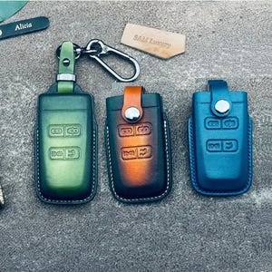 Op de afbeelding: Drie leren sleuteletuis in groen, bruin en blauw. Elke etui heeft een knoopsluiting en reliëf sleutelsymbolen. Een zilveren karabijnhaak en twee kleine leren labels met de namen "Alicia" en "SAM Luxury" zijn ook zichtbaar.