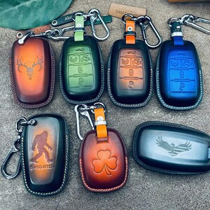 May include: A collection of seven leather key fob covers in various colors including brown, green, orange, blue, and black. Each cover features unique embossed designs such as a deer skull, a sasquatch, a shamrock, and an eagle with a flag.