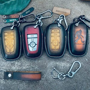 May include: A collection of leather key fob covers in various colors, including red, brown, and black. Some covers feature embossed designs, while others have keychains and metal clasps. One cover has a Bigfoot graphic.