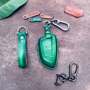 May include: Green leather key fob with a silver key ring and a silver clip. The key fob has a green leather strap with a silver buckle. The key fob is surrounded by four small leather tags with names on them: "Big Goldi", "Karina", "Jeremy", and "Sam Luxury".