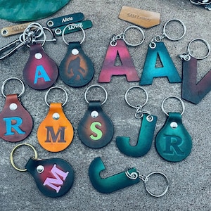 May include: A collection of leather keychains with different letters and designs. The keychains are in various colors, including blue, green, red, orange, and brown. Some of the keychains have a simple letter design, while others have more intricate designs, such as a Bigfoot.