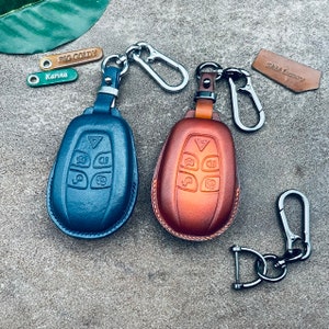 May include: Two leather key fobs with metal key rings. The key fobs are in blue and orange leather and have a design of a car key with buttons. The key rings have a silver finish.