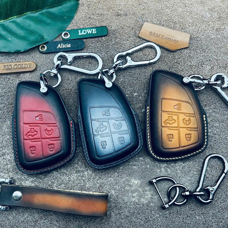 May include: Three leather car key fob covers in red, black, and brown, each featuring embossed button icons and a silver metal carabiner clip. Included are small rectangular leather tags with the text LOWE, Alicia, BIG GOLDY, and SAM Luxury.