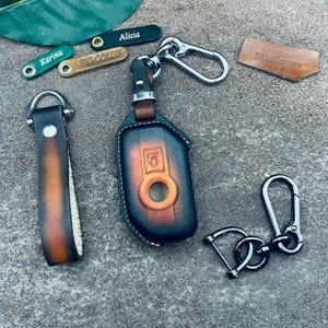 May include: A collection of leather keychains and key holders in various styles. The keychains include personalized tags with names like "Alicia" and "Karina." A key fob case and a key strap are also visible. The color palette includes brown, black, and silver.