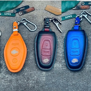 Könnte beinhalten: Eine Auswahl an Leder-Schlüsselanhänger-Hüllen in Orange, Rot und Blau, jeweils mit einem silbernen Karabinerhaken. Die Schlüsselanhänger haben geprägte Autokey-Designs. Kleine Lederanhänger mit Namen wie "Lowe" und "Alicia" sind ebenfalls sichtbar.