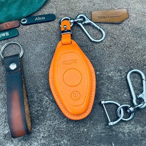 Könnte beinhalten: Ein orangefarbener Autoschlüsselanhänger aus Leder mit silbernen Metallbefestigungen, der eine Autosilhouette und Knopf-Symbole zeigt. Weitere Leder-Schlüsselanhänger und Namensschilder sind ebenfalls sichtbar.