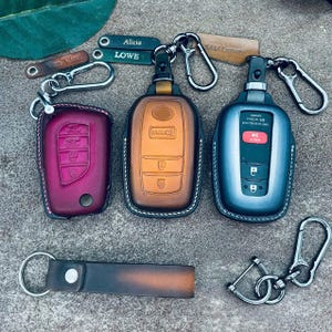 May include: Three leather key fob covers in burgundy, tan, and dark gray, each with a metal carabiner. Two small leather tags are engraved with the names Alicia and Lowe. A separate dark brown leather keychain strap with a metal ring is included.