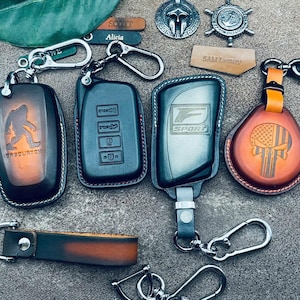 May include: Five leather keychains with different designs. One keychain features a brown leather case with a Sasquatch graphic and the text "Sasquatch" on it. Another keychain has a black leather case with a silver metal key fob with buttons. A third keychain has a grey leather case with a silver metal key fob with the text "F Sport" on it. A fourth keychain has a brown leather case with a skull and American flag graphic. The fifth keychain is a brown leather strap with a silver metal hook.