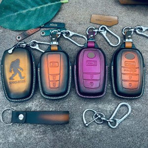 Könnte beinhalten: Eine Sammlung von Leder-Schlüsselanhängern in verschiedenen Farben, darunter Orange, Lila und Schwarz. Ein Anhänger zeigt eine Sasquatch-Grafik mit dem Text "SASQUATCH". Jeder Anhänger hat einen Metallverschluss und einen Schlüsselring. Zusätzliches Lederzubehör ist ebenfalls enthalten.