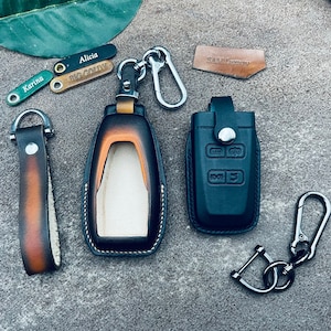 Peut inclure: Deux housses de clés en cuir, l'une marron et l'autre noire, avec un porte-clés argenté et une chaîne de clés argentée. La housse de clé marron a un intérieur blanc et la housse de clé noire a quatre boutons.