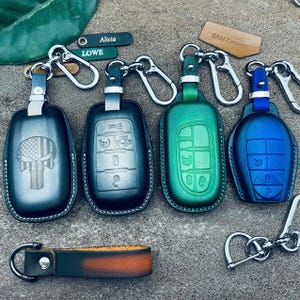 May include: Four leather car key fob covers in black, green, and blue with silver metal clips. One black cover features a Punisher skull and American flag design. Includes small leather tags labeled Alicia, Lowe, and Sam Luxury, plus a leather strap.