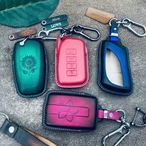 May include: A collection of leather car key fob covers in various colors including green with a sunflower engraving, red, blue, and a pink-to-black gradient with a flower design. Each cover features metal hardware, key rings, and carabiner clips.