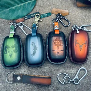 May include: A collection of leather key fob covers in various colors, including green, blue, and brown. Each cover features a unique engraved design, such as a ram skull, eagle, and deer skull. Keychains and metal clasps are also visible.