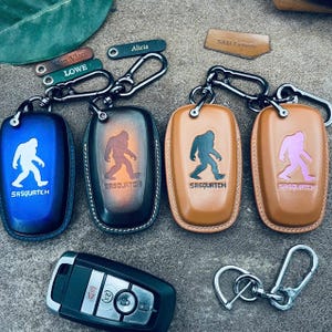May include: Leather key fob cases in blue, brown, and tan, each featuring a Sasquatch silhouette and the word "Sasquatch." Keychains with personalized tags are also visible. A car key and a separate keychain are also included.