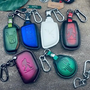 May include: A collection of six leather key fob covers with metal clips. The covers feature various embossed designs including a hand gesture, a dragonfly, a Bigfoot silhouette with the text Sasquatch, and a skull with an American flag pattern.