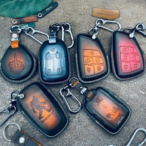 May include: A selection of leather key fob covers in different shapes and colours. Some have embossed designs, including a hand gesture, a Sasquatch, and car key button layouts. Each cover has a metal keyring.