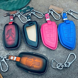 Könnte beinhalten: Leder-Schlüsselanhänger in Rot, Schwarz, Pink und Blau. Einige haben geprägte Designs: einen Sasquatch, ein Auto-Schlüssel-Layout und ein Raumschiff. Jeder Schlüsselanhänger hat einen Metallverschluss zum Befestigen von Schlüsseln.