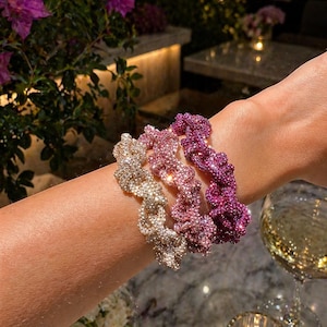 Puede incluir: Tres coleteros brillantes en tonos dorados, rosas y morados se muestran en una muñeca. Los coleteros están cubiertos de pequeños adornos brillantes. El fondo incluye una copa de vino, una vela encendida y flores.