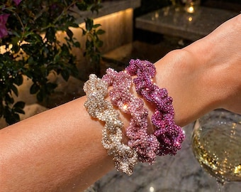 May include: Three sparkling scrunchies in gold, pink, and purple sit on a wrist. The scrunchies are covered in small, glittering embellishments. The background includes a glass of wine, a lit candle, and flowers.
