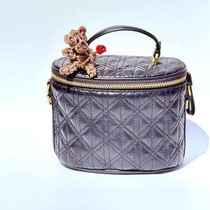 May include: A black quilted vanity case with a gold zipper and handle. A small, beaded teddy bear with a red heart is perched on top. The bag has a diamond pattern.