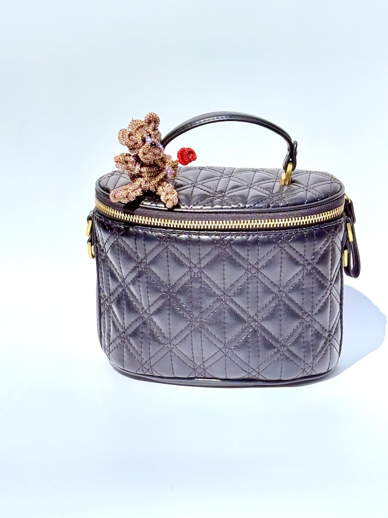 May include: A black quilted vanity case with a gold zipper and handle. A small, beaded teddy bear with a red heart is perched on top. The bag has a diamond pattern.