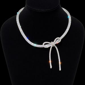 May include: A silver-toned necklace featuring a continuous band of clear, sparkling rhinestones. The necklace is designed with a central bow detail that includes two dangling ends, creating a refined and symmetrical appearance with light-reflecting stones.