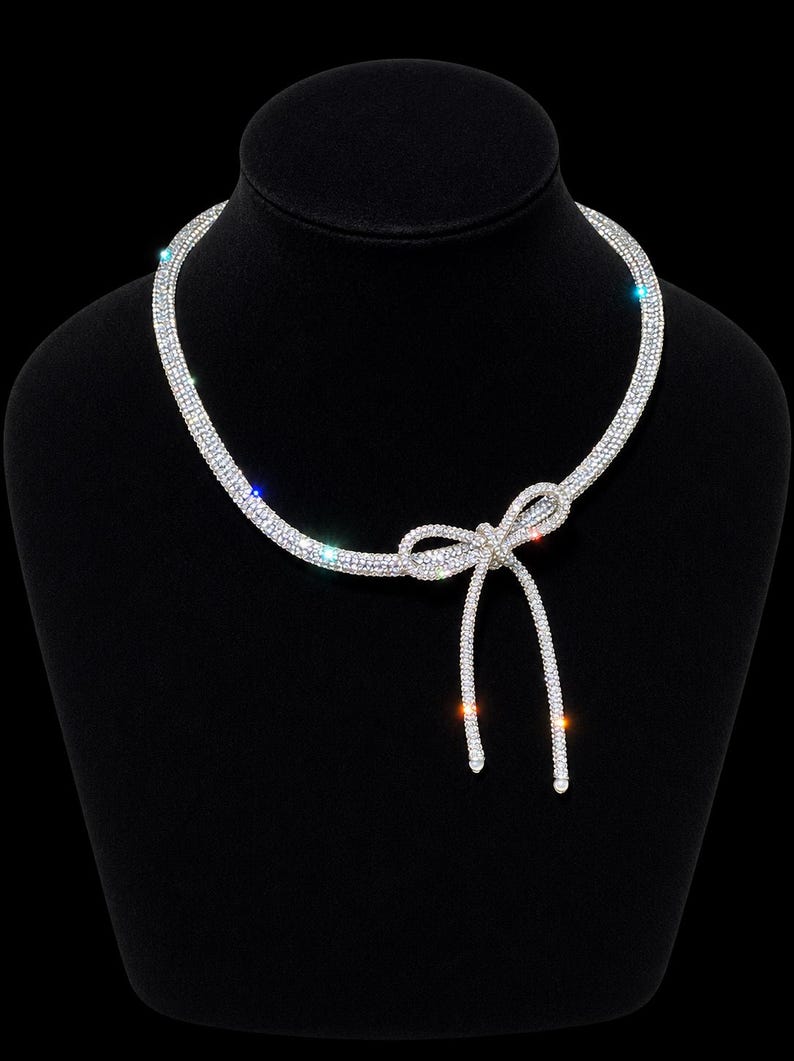 May include: A silver-toned necklace featuring a continuous band of clear, sparkling rhinestones. The necklace is designed with a central bow detail that includes two dangling ends, creating a refined and symmetrical appearance with light-reflecting stones.