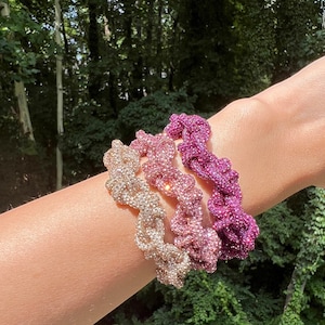 May include: Three beaded bracelets in a gradient of pink, rose gold, and gold. The bracelets are made of small beads that are woven together in a twisted pattern.
