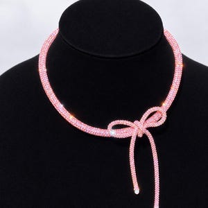 May include: A pink choker necklace featuring a flexible, tubular band covered in small, sparkling crystals. The necklace is tied into a bow at the front, with two dangling ends that each feature a small, round white bead at the very tip.