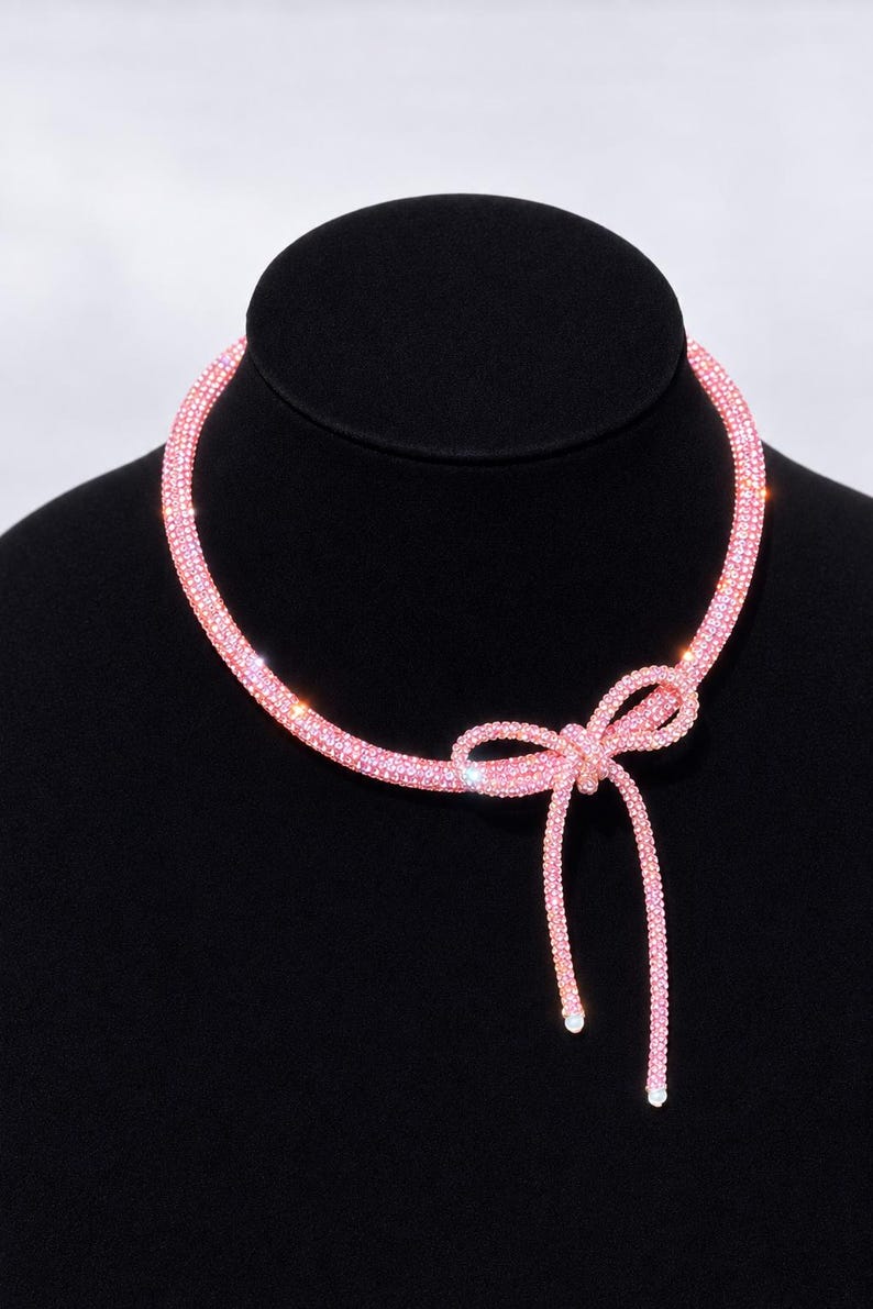 May include: A pink choker necklace featuring a flexible, tubular band covered in small, sparkling crystals. The necklace is tied into a bow at the front, with two dangling ends that each feature a small, round white bead at the very tip.