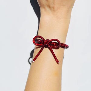 May include: A bracelet featuring a vibrant red, crystal-encrusted bow design. The band is fully covered in small, sparkling red stones, creating a textured, reflective surface that wraps around the wrist with two dangling bow ends.