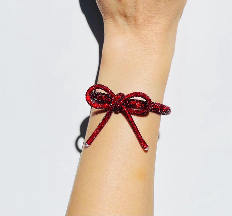 May include: A bracelet featuring a vibrant red, crystal-encrusted bow design. The band is fully covered in small, sparkling red stones, creating a textured, reflective surface that wraps around the wrist with two dangling bow ends.