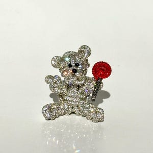 May include: A sparkling, rhinestone-covered teddy bear brooch holding a red lollipop. The bear has black eyes and nose. The brooch is set against a white background.