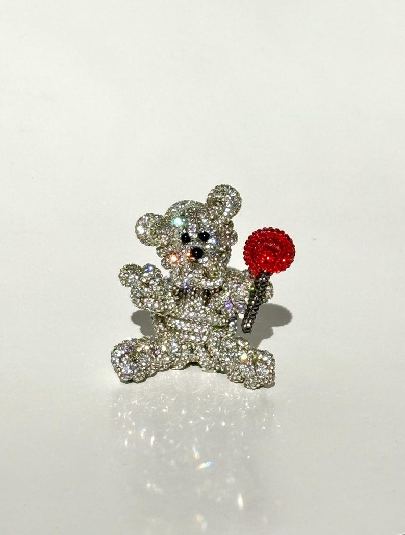 May include: A sparkling, rhinestone-covered teddy bear brooch holding a red lollipop. The bear has black eyes and nose. The brooch is set against a white background.