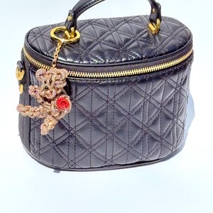 May include: Black quilted vanity case with a gold zipper and handle. A gold-toned charm of a teddy bear with a red rose hangs from the zipper. The bag has a diamond pattern.