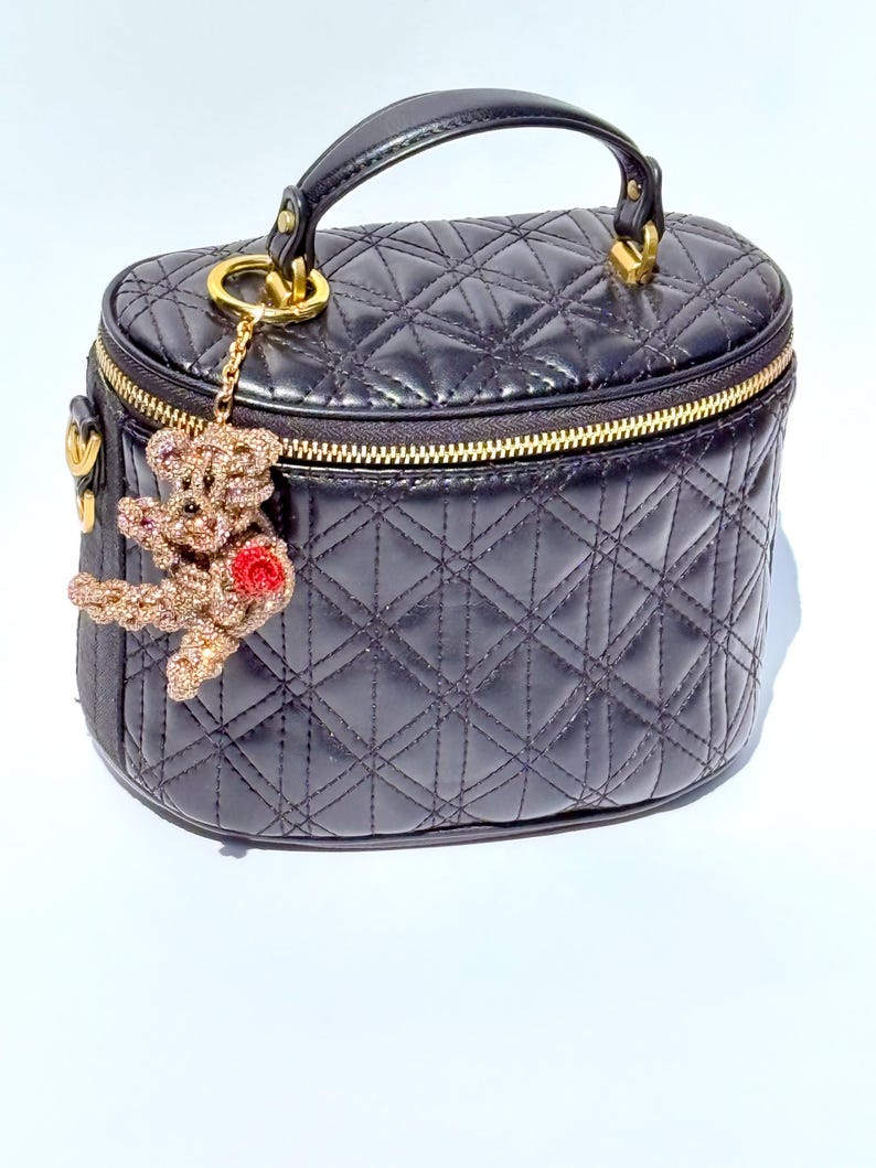 May include: Black quilted vanity case with a gold zipper and handle. A gold-toned charm of a teddy bear with a red rose hangs from the zipper. The bag has a diamond pattern.