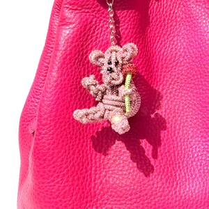 May include: A pink jeweled teddy bear keychain with a red flower, hanging from a silver ring. The teddy bear is covered in small, sparkling crystals. The background is a bright pink leather bag.