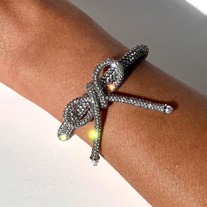 Puede incluir: Un brazalete plateado con un diseño de lazo hecho de brillantes diamantes de imitación.