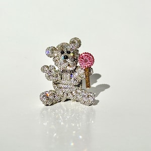 May include: A sparkling, rhinestone-covered teddy bear brooch. The bear is holding a pink lollipop with a gold-colored stick. The brooch is set against a white background, creating a glamorous and eye-catching accessory.