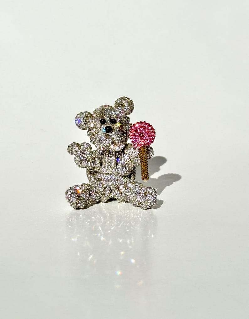 May include: A sparkling, rhinestone-covered teddy bear brooch. The bear is holding a pink lollipop with a gold-colored stick. The brooch is set against a white background, creating a glamorous and eye-catching accessory.