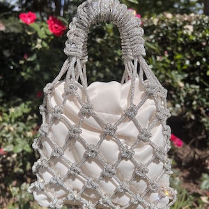 May include: A white satin bag with a diamond-encrusted net design. The bag has a top handle and is made of a delicate, intricate material.