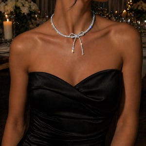 May include: A silver-toned choker necklace featuring a central bow design encrusted with clear, sparkling stones. The necklace sits closely around the neck, with the bow detail positioned at the centre, creating a refined and polished appearance.