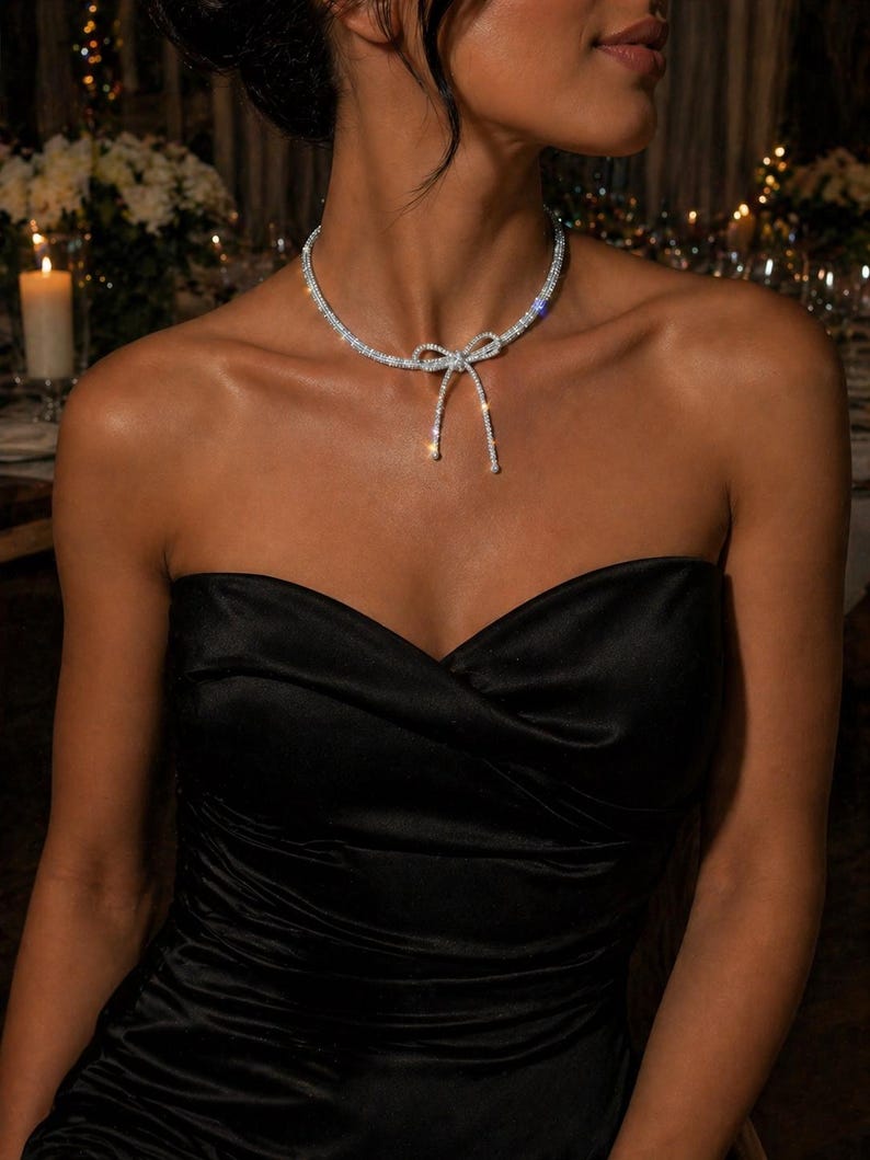 May include: A silver-toned choker necklace featuring a central bow design encrusted with clear, sparkling stones. The necklace sits closely around the neck, with the bow detail positioned at the centre, creating a refined and polished appearance.